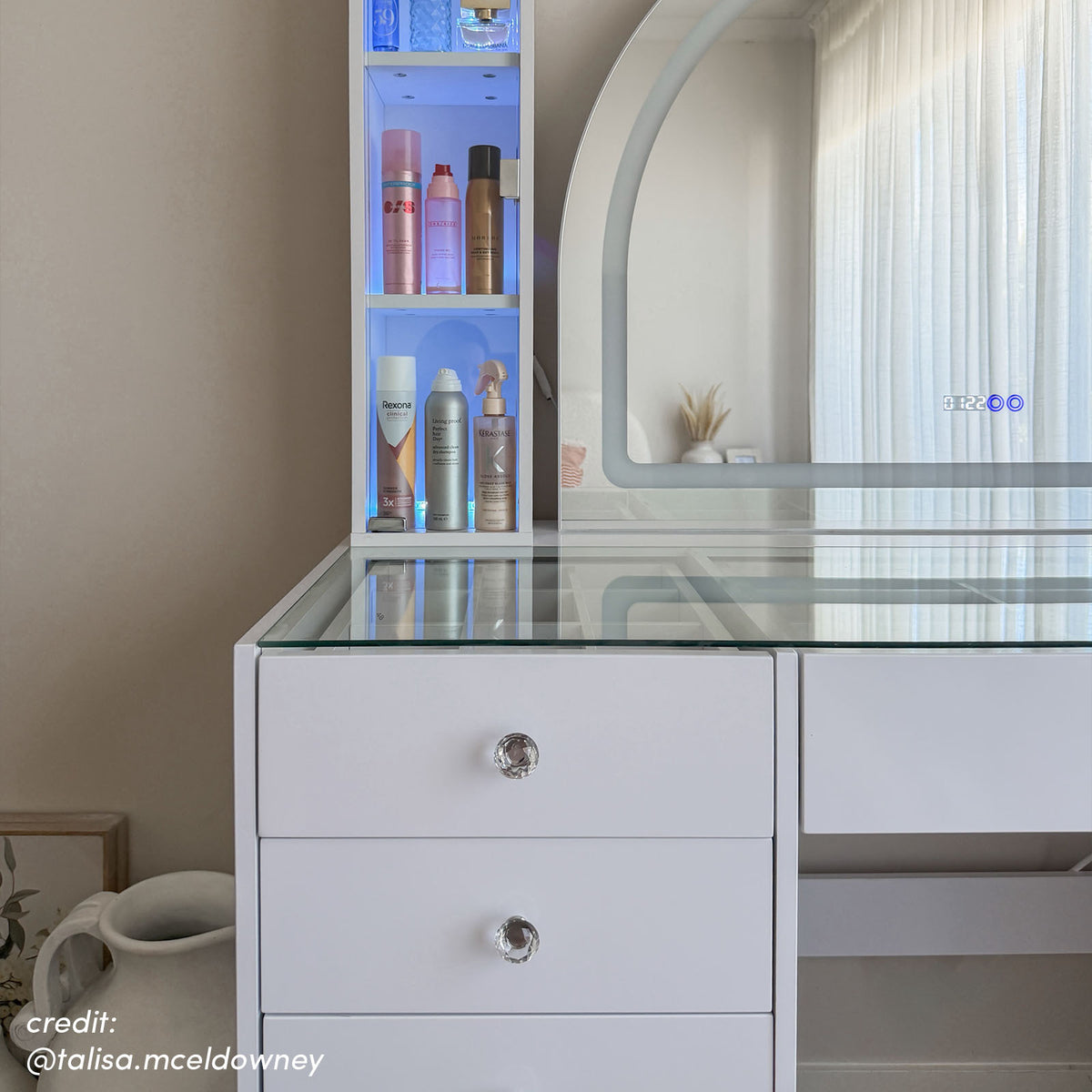 White vanity table with glass top, crystal drawer knobs, arched LED mirror and illuminated side shelving displaying beauty
