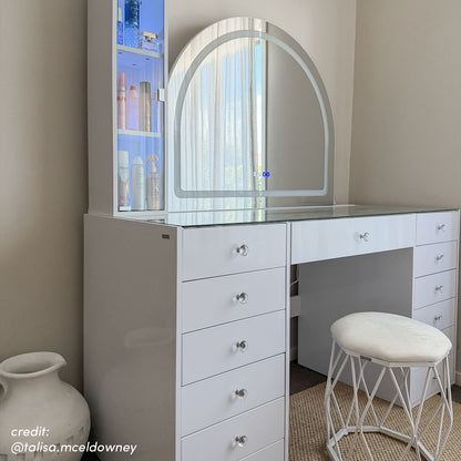 White vanity table with arched LED mirror, glass top, crystal drawer knobs, and matching stool in neutral room