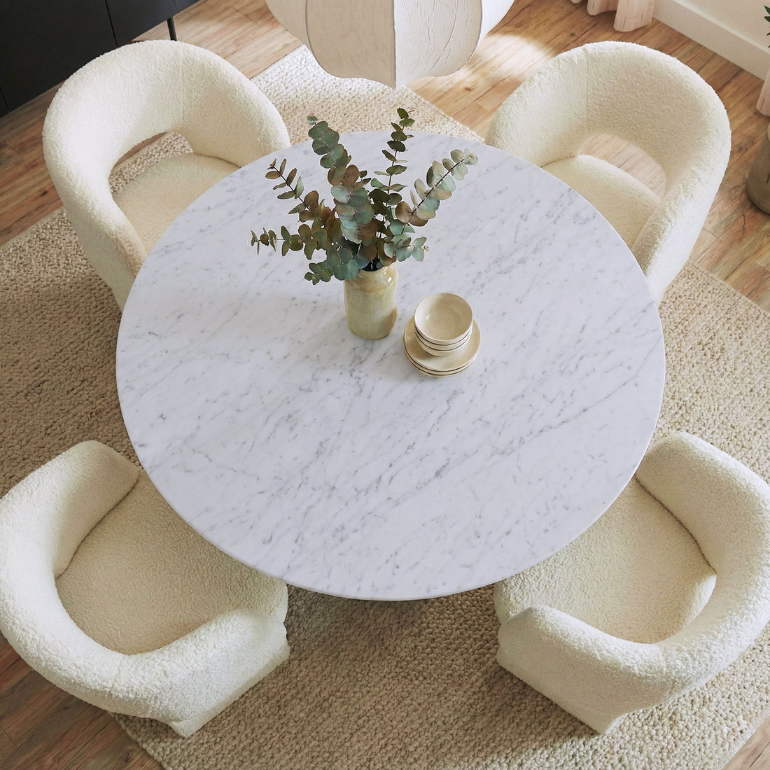 Dina 120cm Round Dining Table in Real Carrara Marble with blue-grey veining, ceramic vase with eucalyptus, light-beige bowls on table, surrounded by four boucle fabric chairs on a natural-fiber rug.