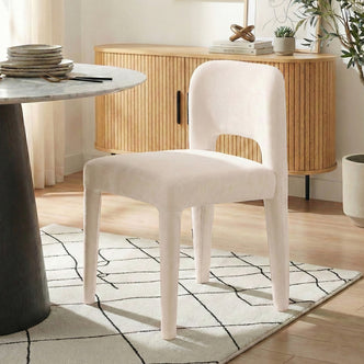 Edris upholstered dining chair in oat fabric with curved backrest, four tapered legs, and cut-out detail, shown beside a
