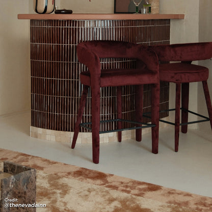 Eisha Velvet Dining Chair in Dark Plum, curved backrest, padded seat, tapered legs with metal footrest, shown at bar height