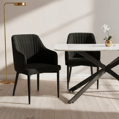 Elly Curved Velvet Dining Chairs Set of 2 in Black, featuring plush upholstery, curved high-back with armrests, and tapered legs, positioned around a marble-top table with a gold vase and orchid.