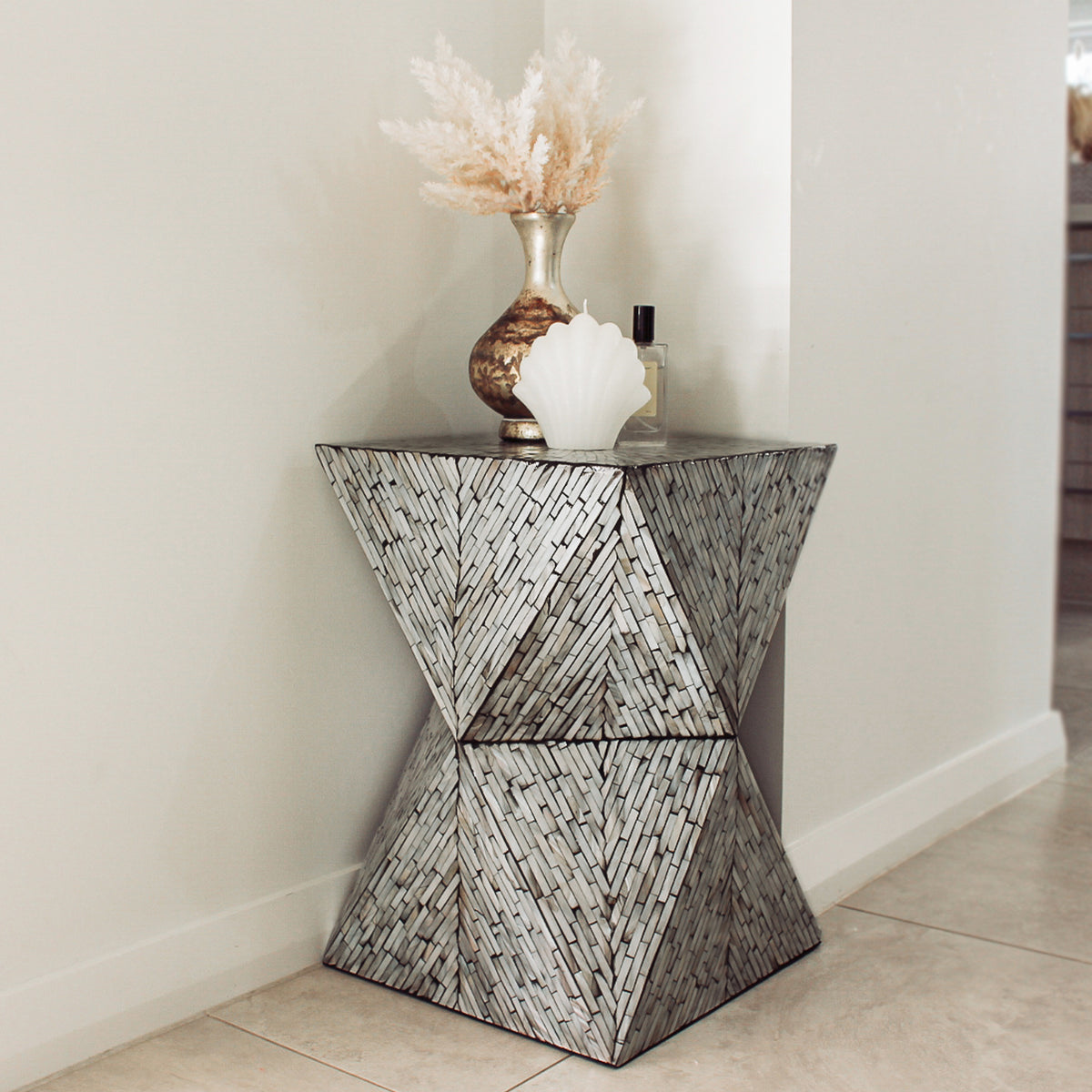 Enki 35cm Mother of Pearl Inlay Side Table in Beige & Black, featuring a geometric hourglass shape with a silvery mosaic finish, topped with a metallic vase, candle, and perfume bottle in a tiled corner.