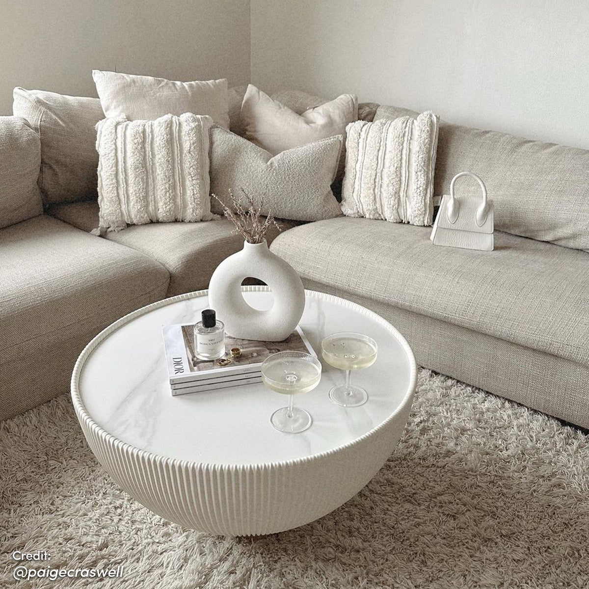 Glalie 70cm Sintered Stone Coffee Table Cream White with ribbed base, set with coupe glasses, ceramic vase, and magazines, complements an L-shaped beige sectional and shaggy rug in a neutral-toned living room.