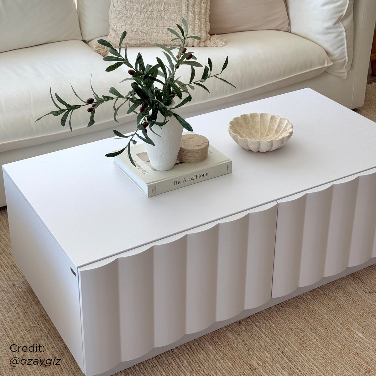 Hamish 120cm Coffee Table with Storage featuring a modern wave design, visible drawers, and a spacious top, complemented by indoor decor elements like a plant in a vase on the table.