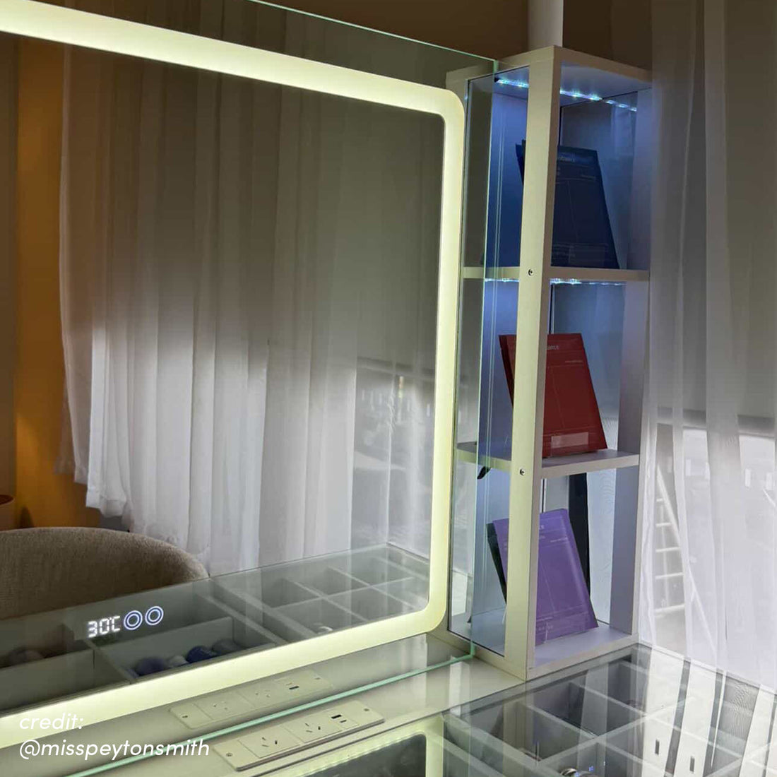 LED vanity mirror with digital display and glass-fronted side cabinet with illuminated shelves holding triangular boxes