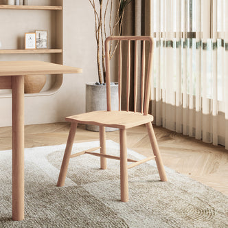 Leigh Solid Oak High-back Dining Chair Natural Set of 2, features a spindle-back design, positioned beside a matching table on a textured rug, with sheer curtains and a potted plant enhancing the room's ambiance.