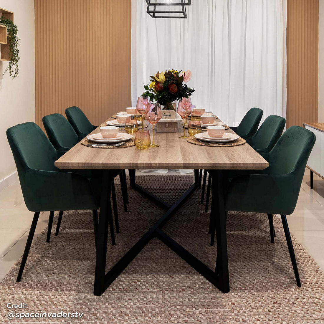 Raynie Velvet Dining Chair Set of 2 in Deep Green, featuring plush velvet upholstery and slender black legs, surrounding a wood-grain table with pink-tinted glassware and a central floral arrangement.