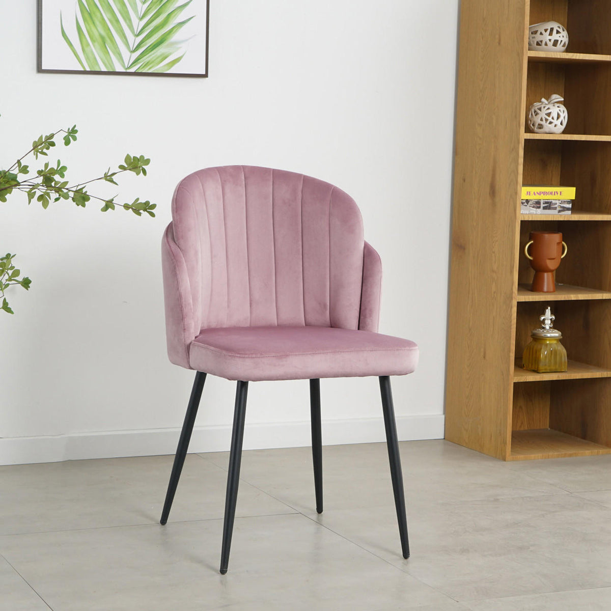 Scarlett Velvet Dining Chair Set of 2 in Mauve Pink, featuring blush-pink velvet upholstery with vertical channel stitching, black tapered metal legs, on a light gray floor, framed by a plant and shelving unit.