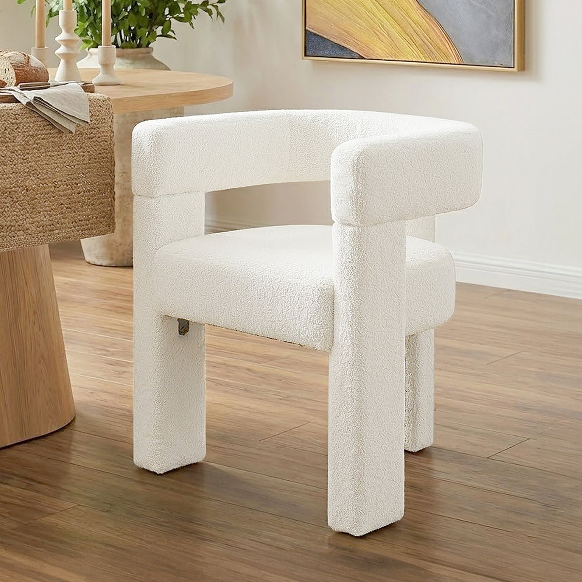 Utah Bouclé Curved 3 Legged Accent Chair in Cream, featuring rounded backrest, creamy-white boucle fabric, and three thick legs, placed on hardwood flooring near a wooden table and abstract art.