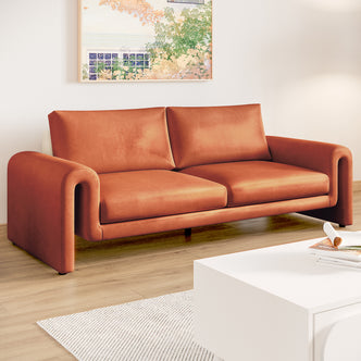 Celyn 3 seater velvet upholstered sofa in burnt orange with curved armrests, shown in a living room with light wood flooring