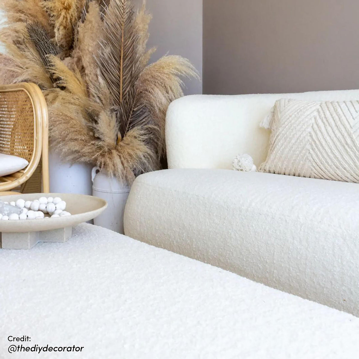 Jamie 110cm Boucle Fabric Ottoman in Cream White, a low, textured piece in a cozy boho setting, complemented by a matching sofa, textured pillow, and sand-colored bowl with wooden beads.