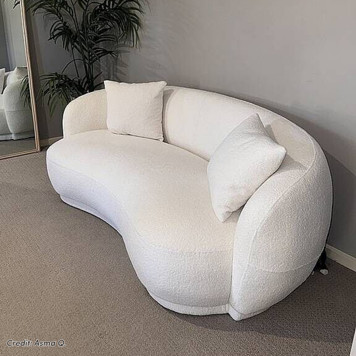A curved white boucle sofa with two matching throw pillows, set against a grey wall on carpet with a tall mirror and plant in the background.