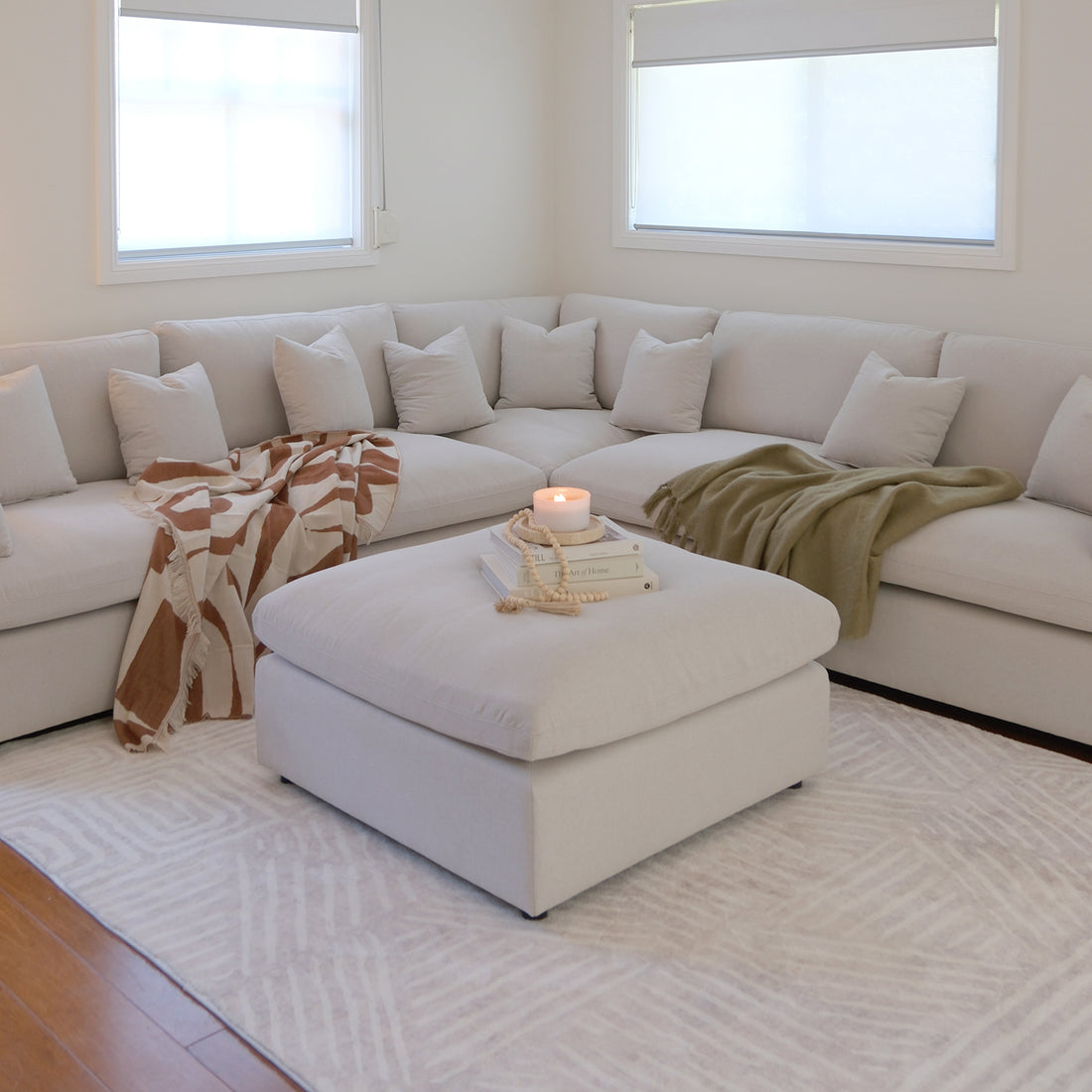 Millie 6-Seater Fabric Corner Chaise Sofa in warm light grey, featuring plush cushions and modular design, elegantly styled with a white pillow and candle-topped ottoman for a cozy living space.
