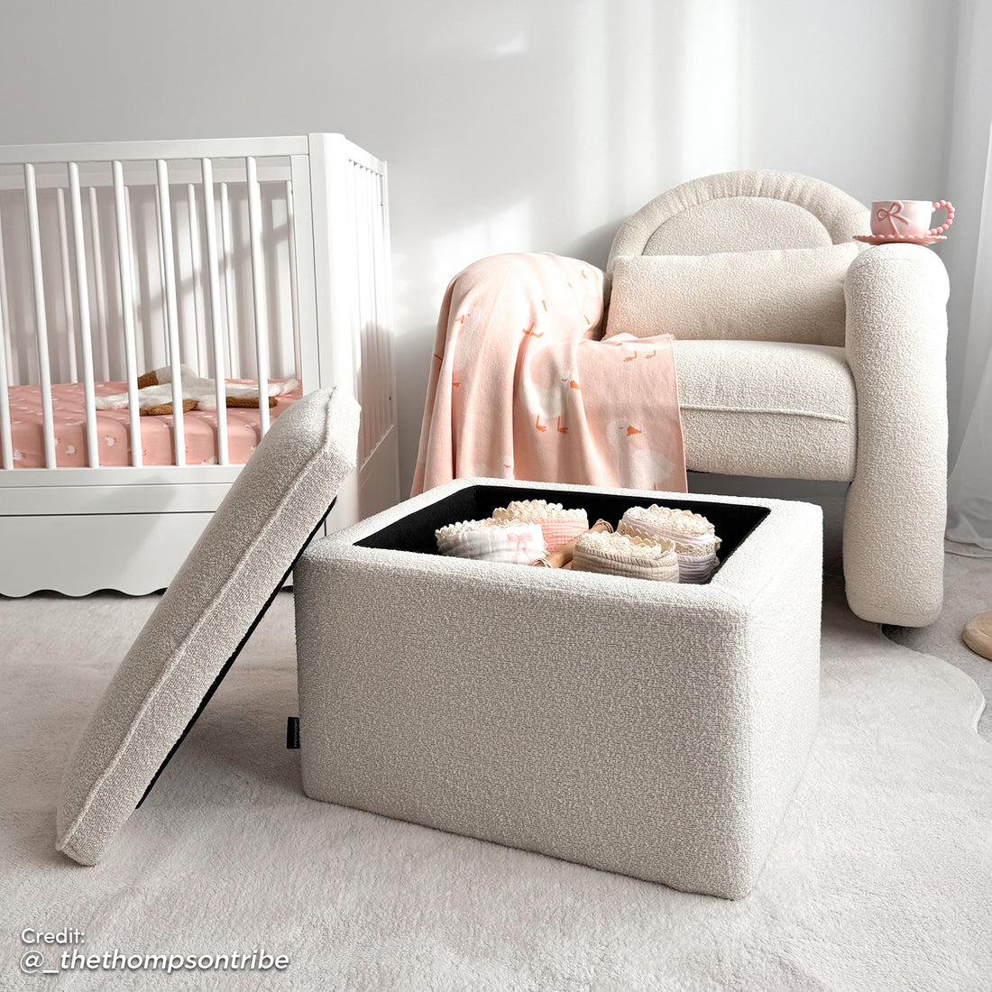 Cream bouclé ottoman with hinged lid open, revealing folded baby clothes inside, beside matching rocking armchair and white