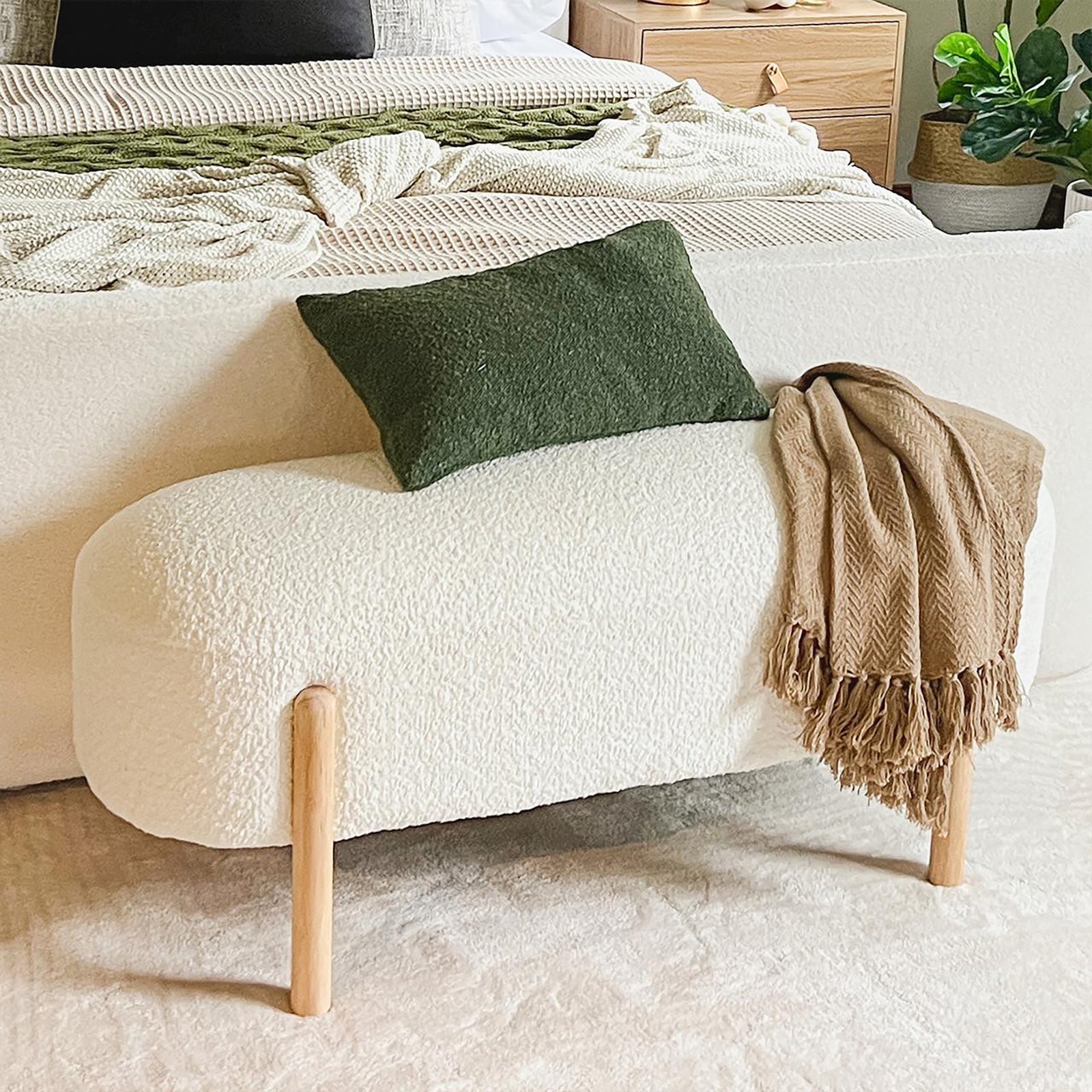 Suellen 108cm Boucle Fabric Ottoman Bench - Cream White, showcasing a plush boucle texture, wooden legs, and tassels, surrounded by a brown blanket and plants in pots, perfect for stylish, versatile seating.