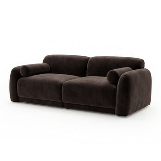 Dark brown velvet sofa with rounded armrests and cylindrical bolster pillows on white background