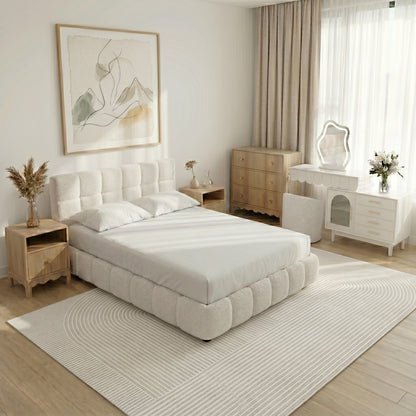 A modern bedroom featuring a tufted cream bouclé bed, light wood bedside tables, a white vanity with a wavy mirror, and a linear-patterned rug.