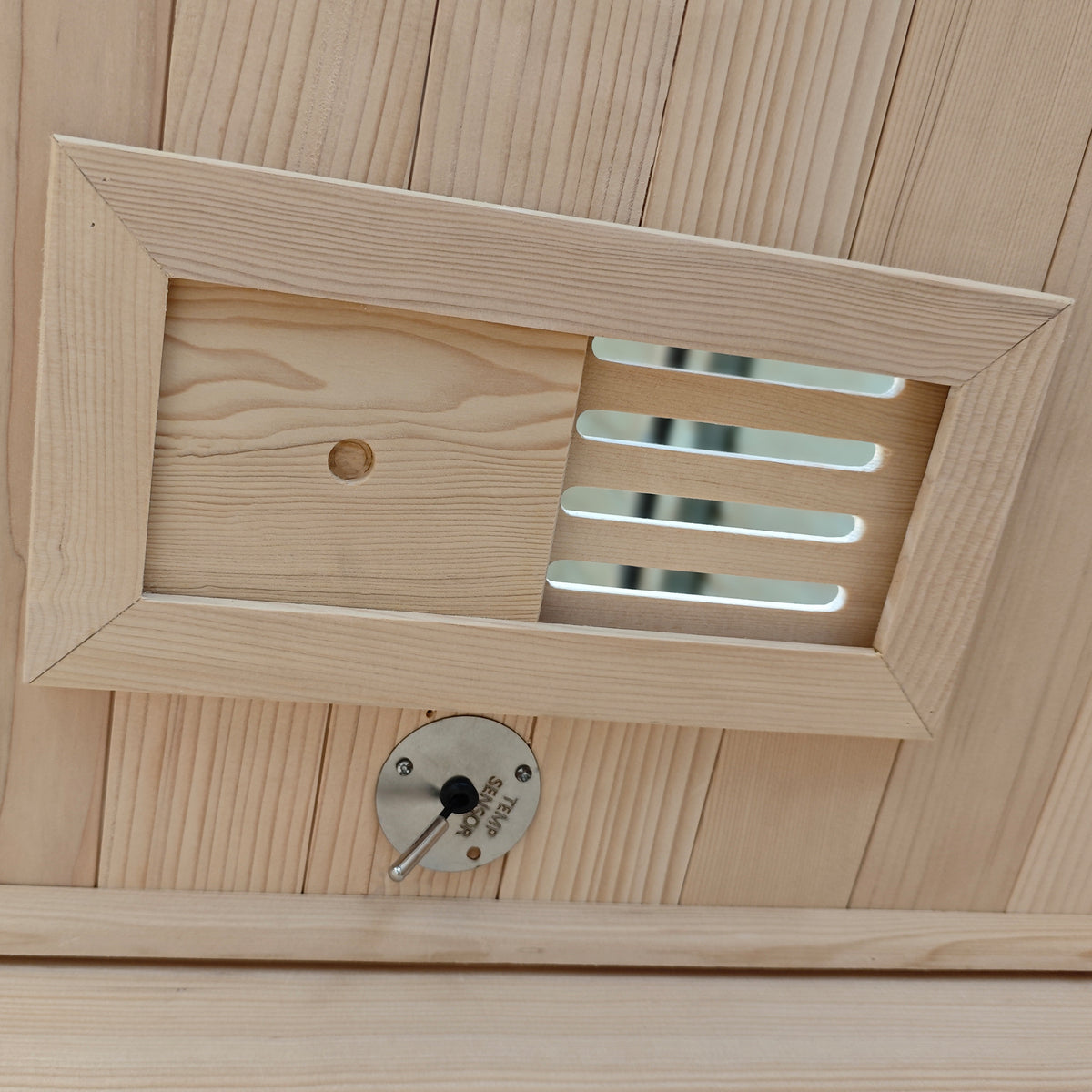 Wooden vent cover with a round metal control dial on a wood-paneled surface.