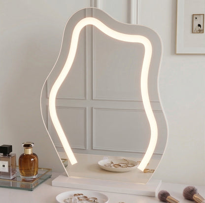 Lifestyle shot of wavy LED-lit vanity mirror on tabletop with makeup brushes, perfume bottles, and jewelry in background