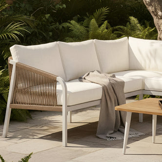 Outdoor lounge with Vernon Sofa Cushion Covers in Cream White on a curved sectional. Features woven rope frame and textured gray throw. Lush foliage backdrop enhances the tranquil setting.