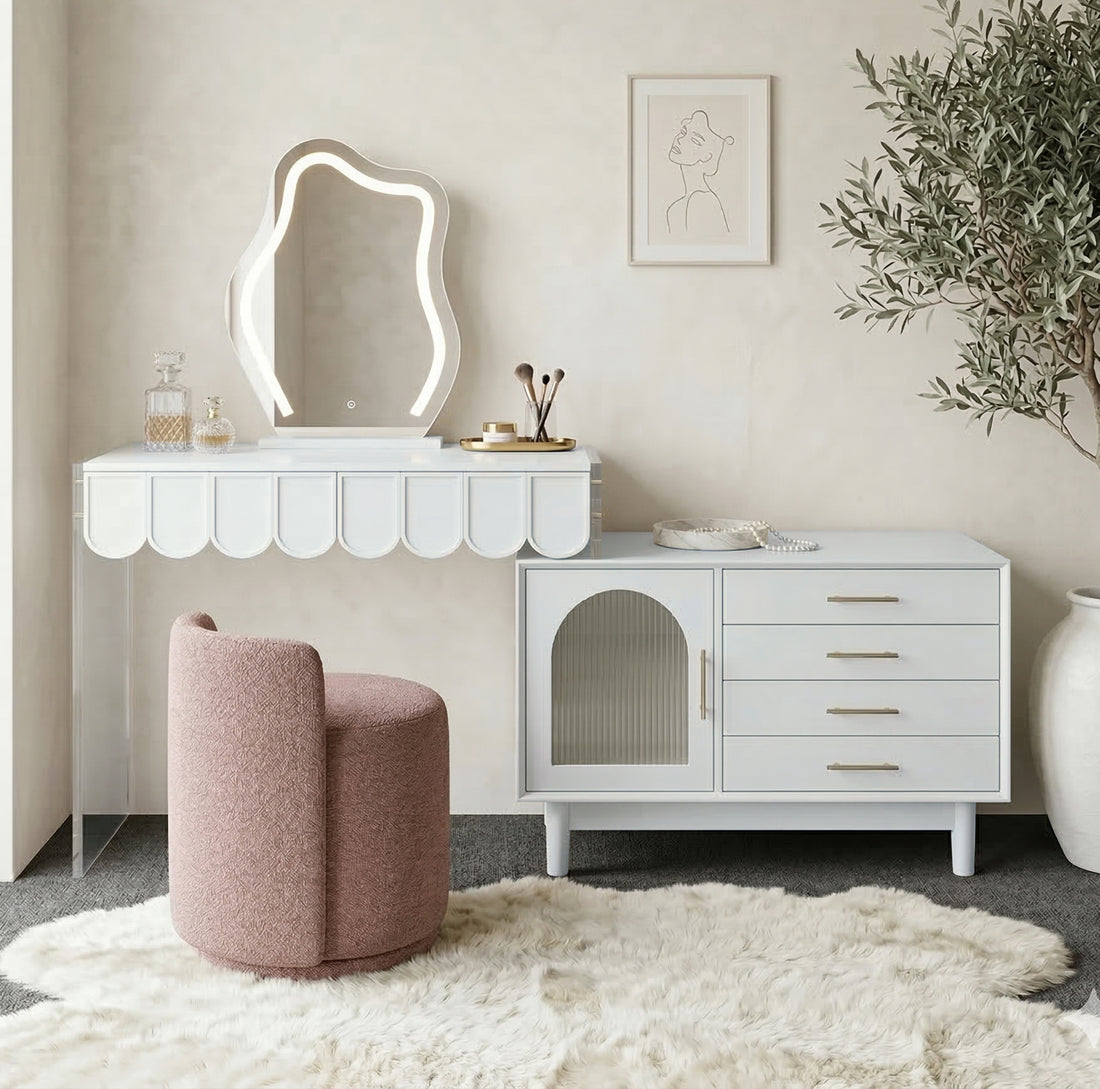 Lifestyle image of the white vanity with scalloped detailing, wavy LED-lit mirror, side cabinet, and blush pink chair in a styled room