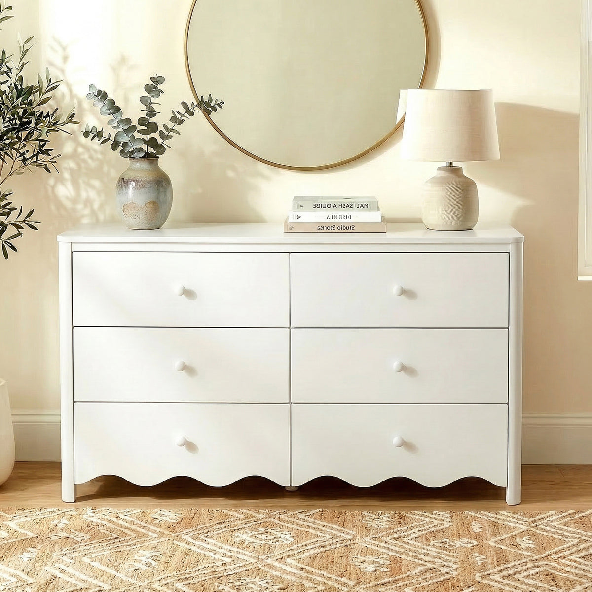White dresser in a softly lit room with vase, books, lamp, and mirror