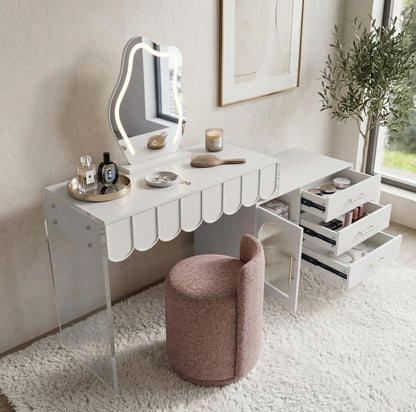 Lifestyle shot of the white scalloped-edge vanity with backlit mirror and pink stool on a textured rug near window and plant
