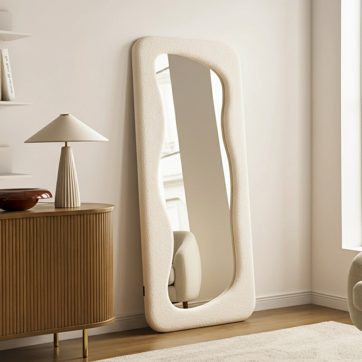 Sabrina 180cm Curved Full Length Mirror in Beige features a padded, undulating frame against a pale wall, enhancing a minimalist living room corner with a serene, contemporary feel.