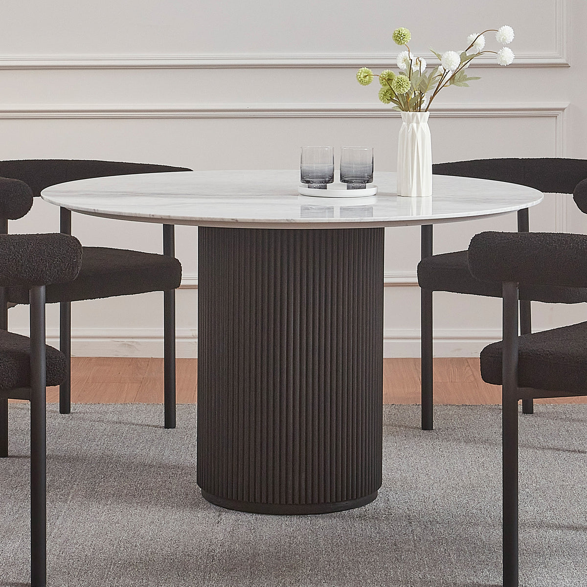 Gracie 120cm Round Marble Dining Table in Black features a white marble top on a black fluted base, surrounded by four plush black chairs, complemented by a vase and glasses on top.