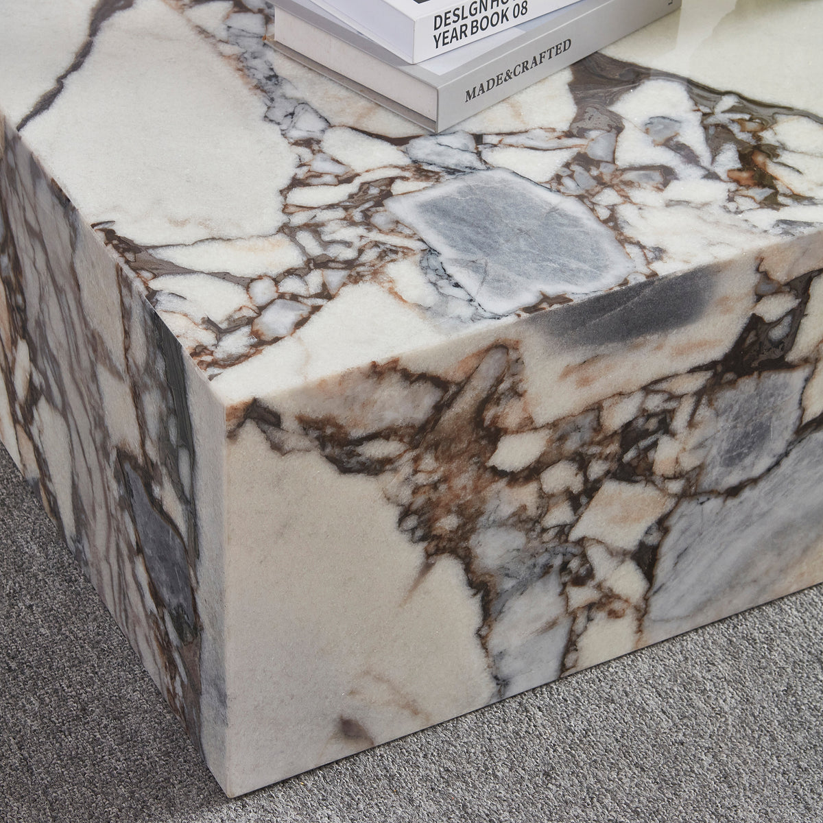 The Solene 100cm Coffee Table in Blue Galaxy features a white marble surface with striking brown and gray veining, topped with hardcover books, set on a low-pile grey carpet.