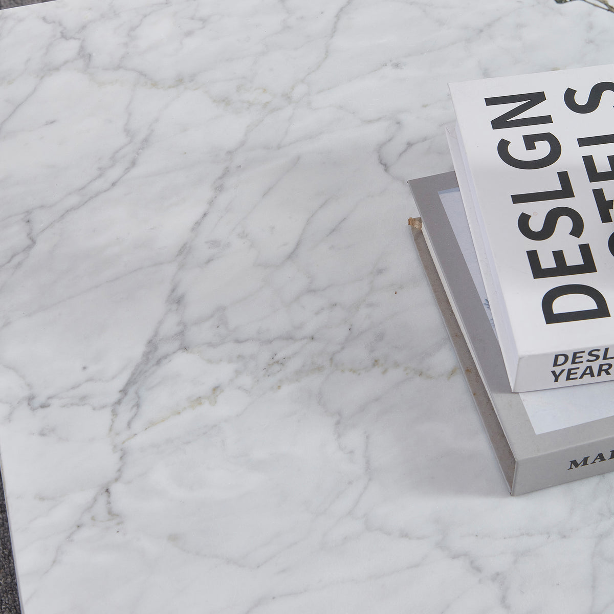 Solene 100cm Coffee Table in Carrara on a white marble surface, featuring a small stack of hardcover books. The table showcases its elegant, authentic marble veining and sleek, low-profile design.
