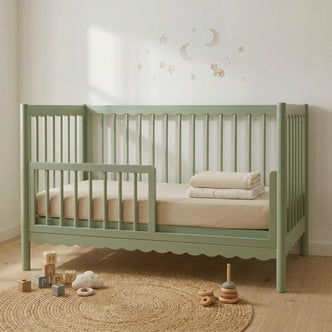 A sage green toddler bed featuring a decorative scalloped trim along the top and bottom rails, set in a neutral-toned nursery with a woven rug and baby toys.