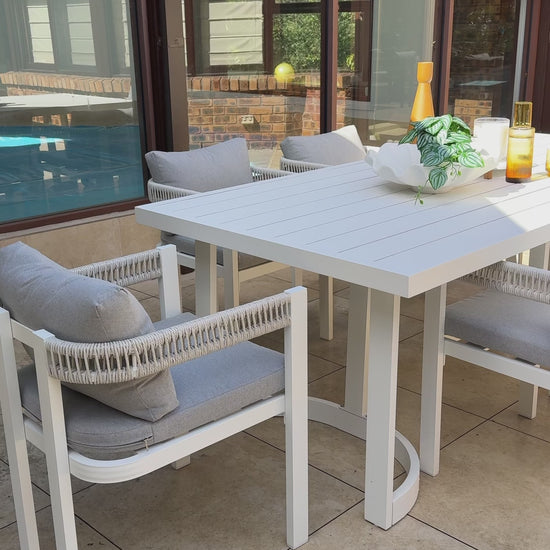 
A wide shot of a modern outdoor dining set. The set includes a large, white rectangular table with a unique half-circle base and multiple chairs. The chairs have white frames, thick gray cushions, and intricate white rope detailing on the backrests. The table is styled with a white bowl of greenery, a yellow vase, and two glasses with a golden-hued drink. The furniture is set on a tiled patio, with a swimming pool visible in the background.