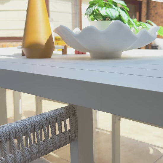 
A close-up of a white, modern outdoor dining table decorated with a gold vase and a wavy white bowl. The camera pans slowly to a chair with a white frame and thick, woven ropes forming the backrest. The seat and back cushions are a comfortable gray, highlighting the elegant design of the outdoor furniture set. The background shows a patio with large windows and a covered seating area.